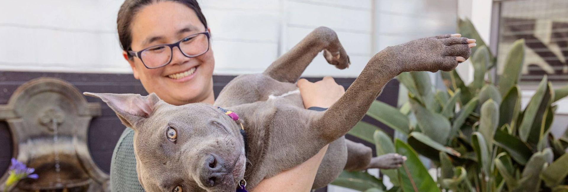 Woman in glasses standing outside with plants surrounding her, holding a grey dog upside as it smiles at the camera.